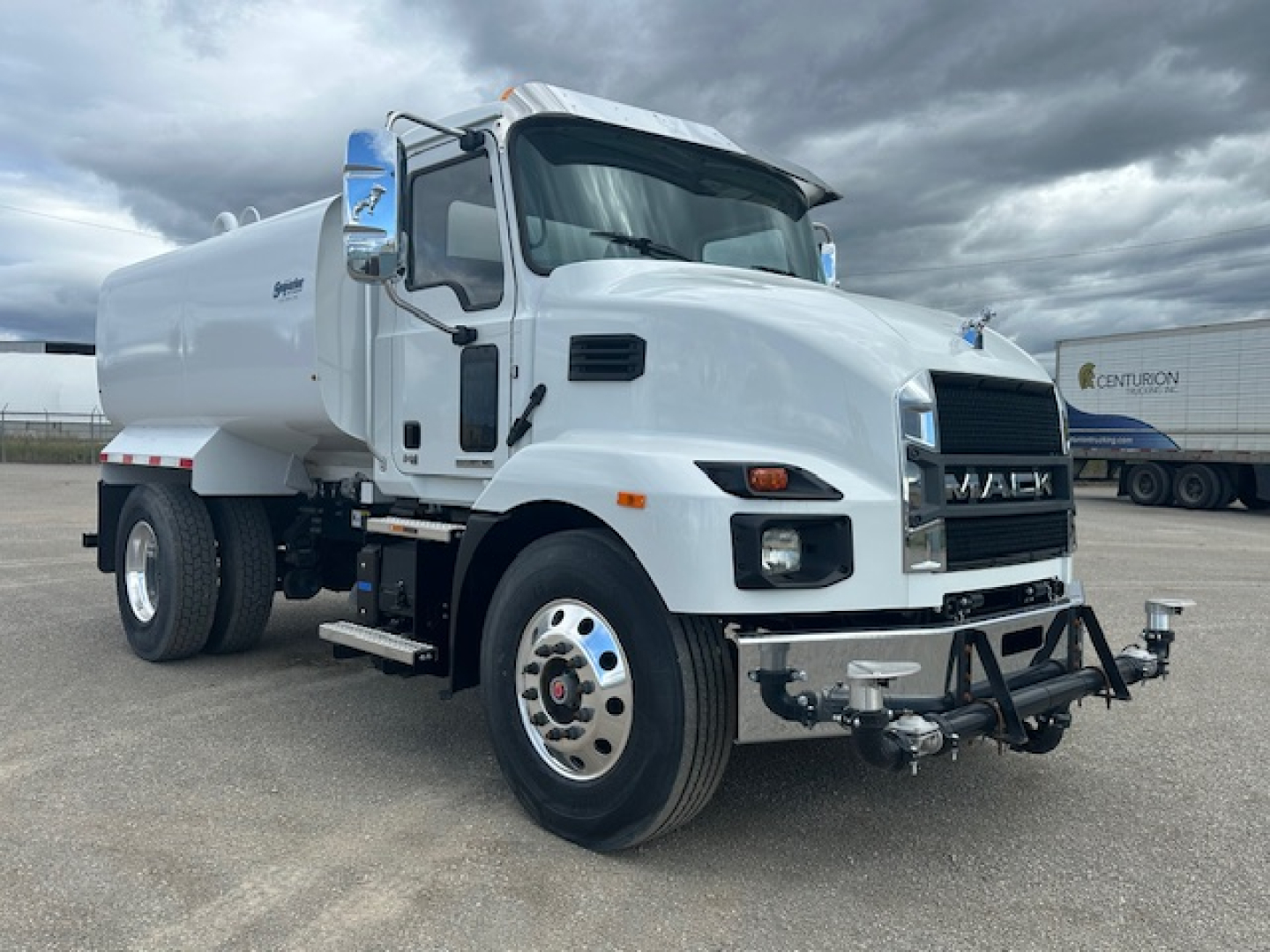Front-left angle of a white 2025 Mack MD7 dump truck with water tank, showing chrome grille, side mirrors, and bumper with towing hardware.