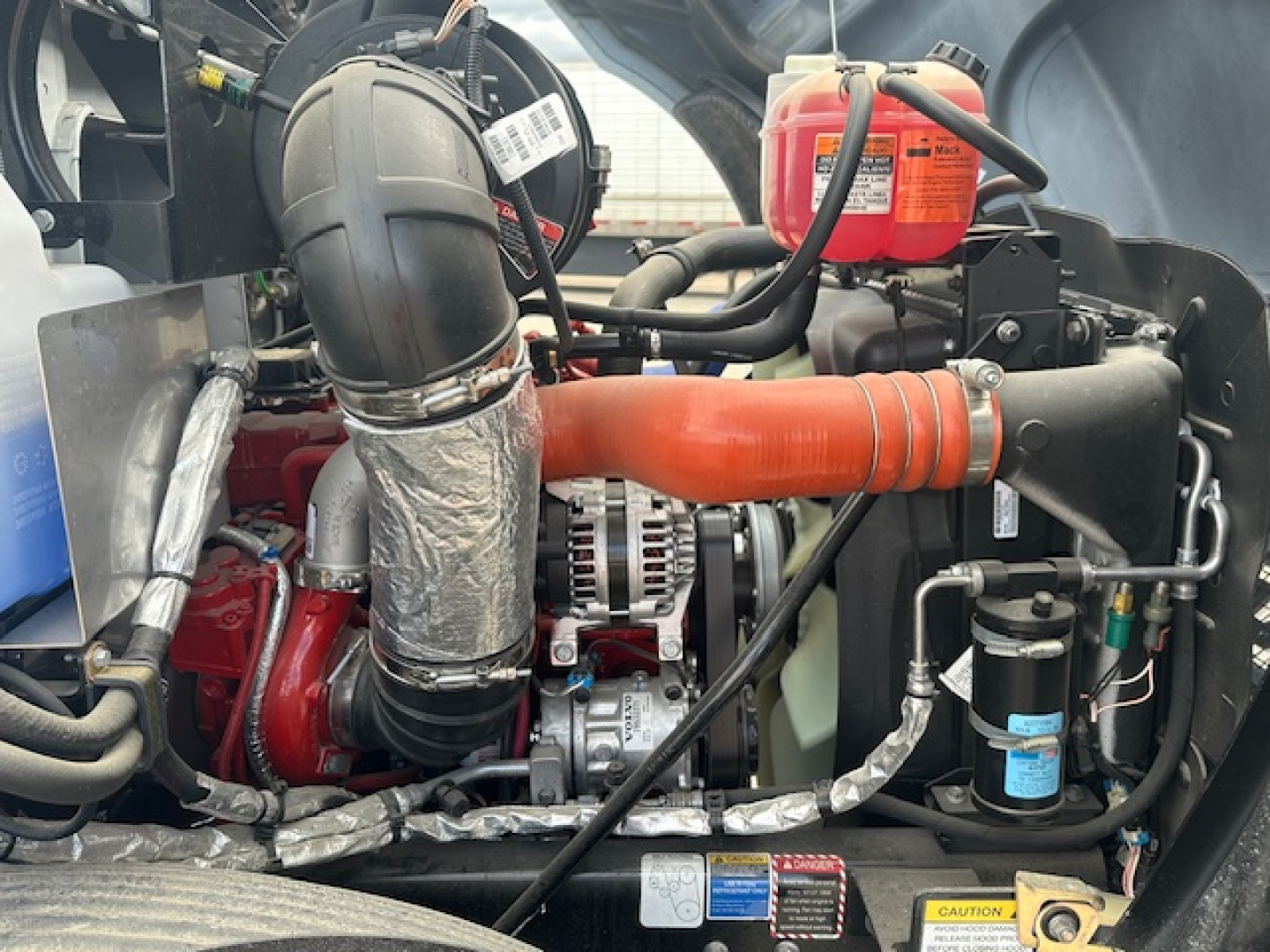 Mack MD7 engine bay showing red intercooler hose, alternator, coolant reservoir, and various hoses and wiring.