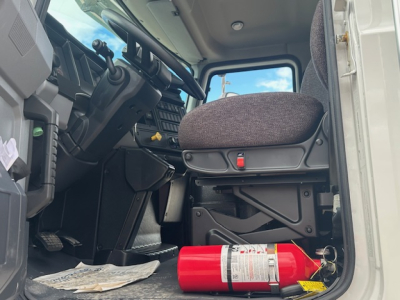 Truck cab interior showing driver seat and steering wheel of a Mack MD7 with a fire extinguisher on the floor.