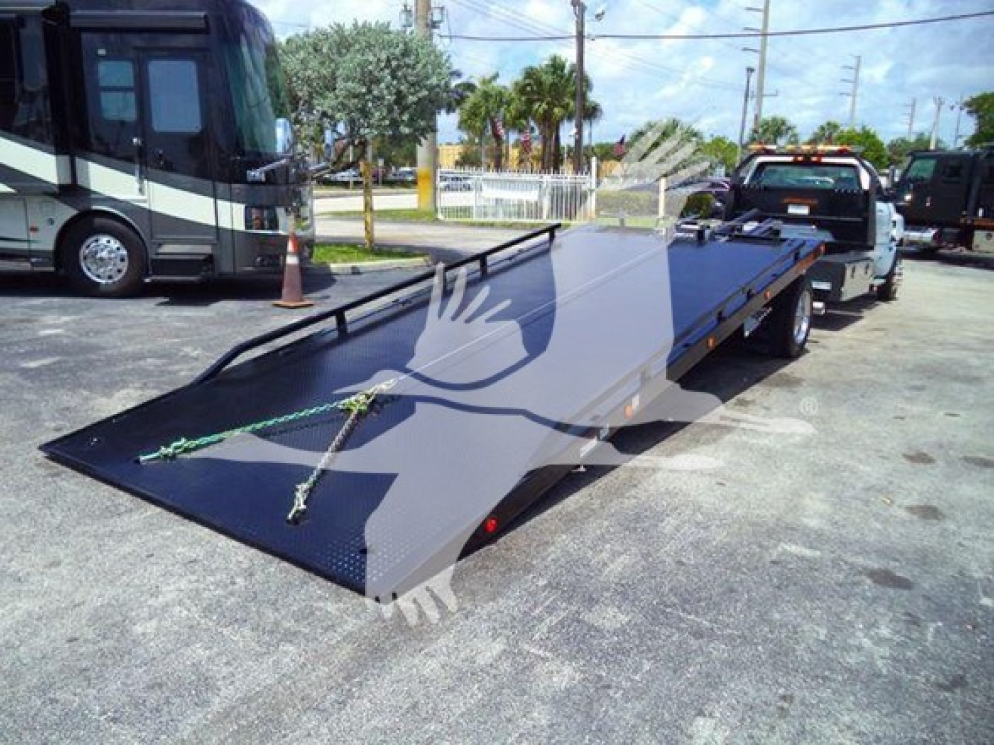 Truck with loaded car hauler/rollback bed in a dealership lot showing a flatbed trailer in the foreground and a tow truck in the background.