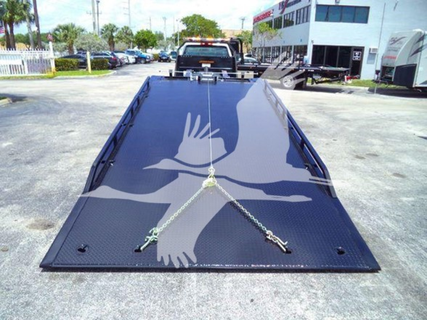 Truck bed of a new 2023 Chevrolet Silverado 6500HD rollback truck showing a metallic black diamond plate surface with chains at the ramp and a bird silhouette reflection.