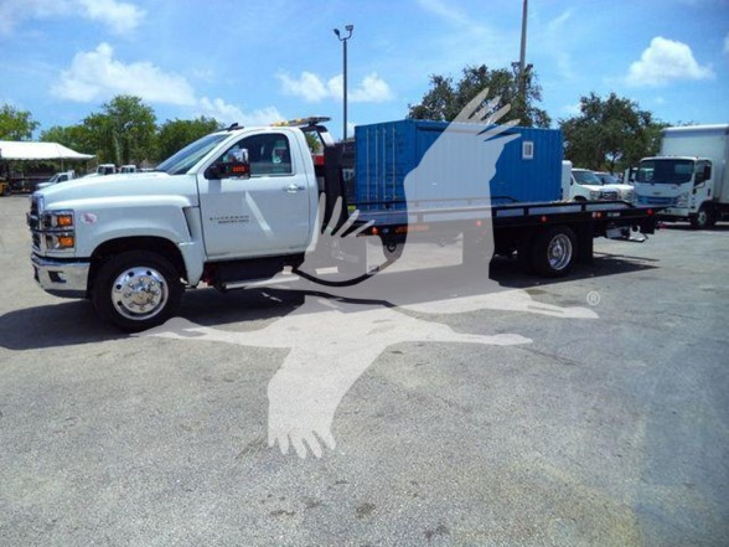 Truck cab with flatbed hauling a blue equipment container in a dealership lot. No visible text legible on signs.