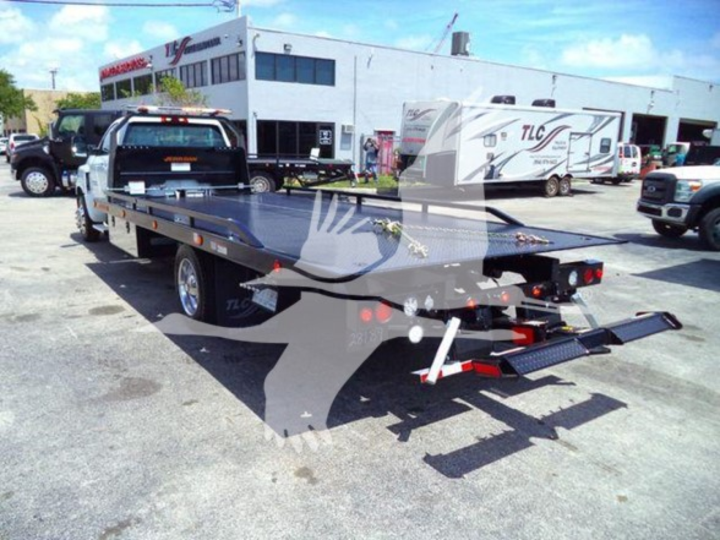 Truck bed showing a flatbed hauler with a Chevrolet Silverado chassis, rear view, loading ramps, and dealership lot in background.