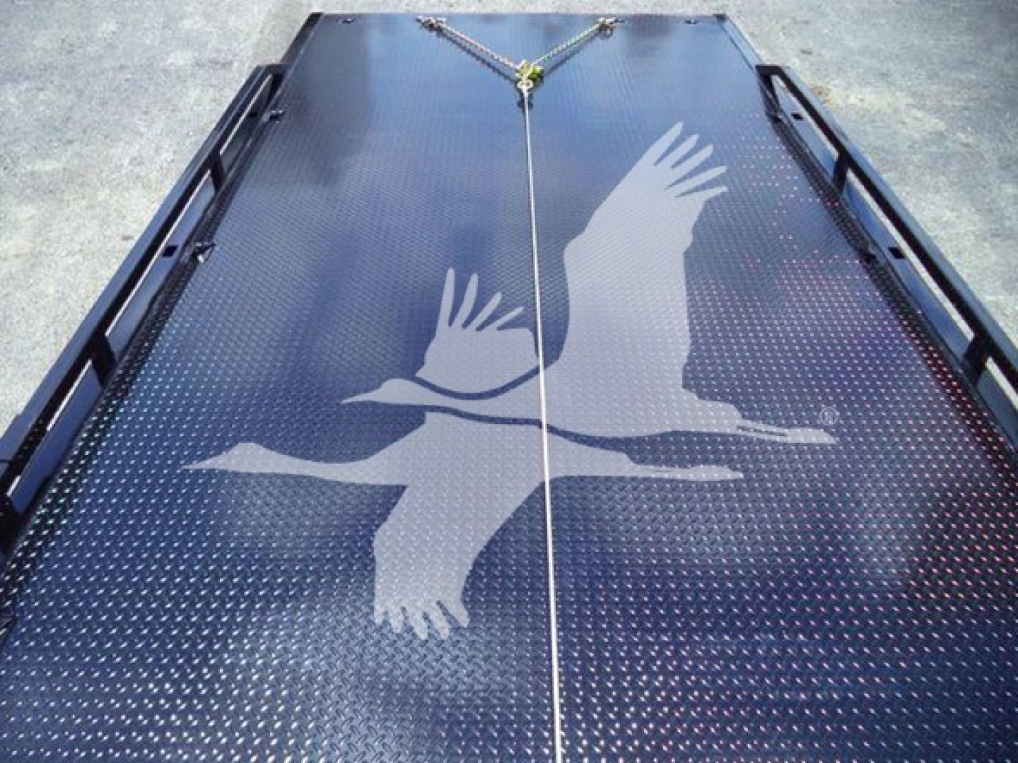 Flatbed trailer/rollback bed with perforated surface and a large white bird graphic; upper center has a tie-down ring and strap. No visible text except the graphic.