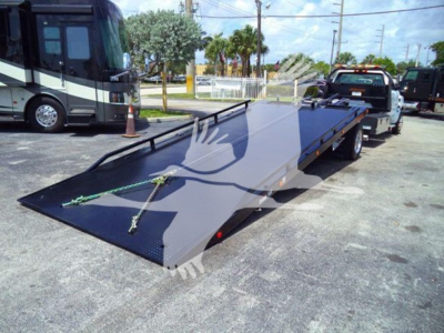 Truck with loaded car hauler/rollback bed in a dealership lot showing a flatbed trailer in the foreground and a tow truck in the background.