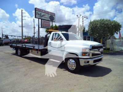 Truck flatbed tow truck in white with long wheelbase and chrome bumper, Silverado 6500HD badge not visible, dealership sign in background.