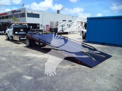 Truck with a long flatbed rollback trailer in a dealership lot; background shows buildings and a blue shipping container.