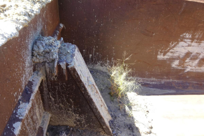 Dump truck bed showing reinforced steel edge and dirt/grime buildup around the lower corner, with a small plant growing near the edge.
