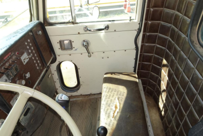 Truck cab interior showing white door with metal handle, window crank, and a brown quilted seat cover; dashboard with gauges and switches visible on left.