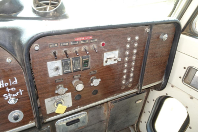 Peterbilt truck dash panel with woodgrain guard, multiple toggle switches, and controls; control cluster on left with labeled switches and a key switch, interior dash shows wear and a yellow tag. 