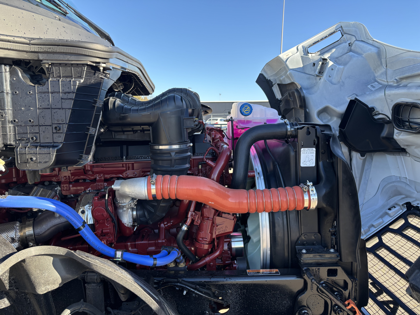 Engine bay view of a 2027 Mack PR64T showing engine components, hoses, radiator, and intake with orange coolant hose and blue hose.