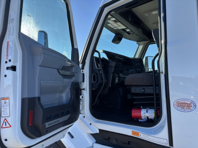 Mack PR64T cab interior view from open driver's door showing dashboard, steering wheel, and seat; door panel with storage pocket and 'MACK' badge visible.