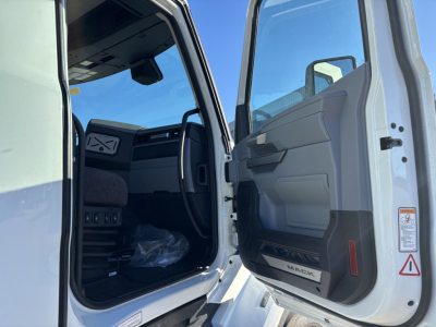 Truck cab interior visible through open driver door of a white Mack PR64T. Dashboard, steering wheel, and door panel with 'MACK' logo on door sill.