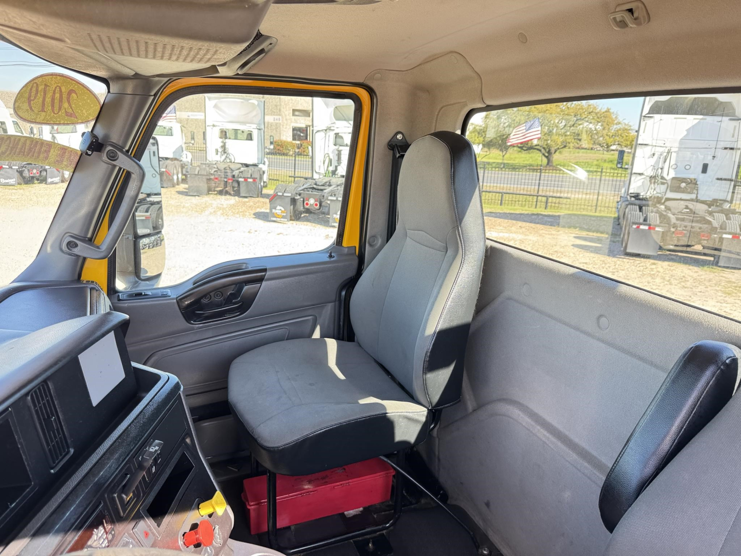 Truck interior showing driver's seat and passenger seat with gray upholstery, yellow door frame, instrument panel, and side window; some red toolbox under seat; exterior lot with other trucks visible through windows.