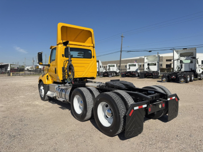 Truck chassis with raised cab in a yard, rear view showing dual rear axles and fifth wheel, yellow cab, no visible branding text