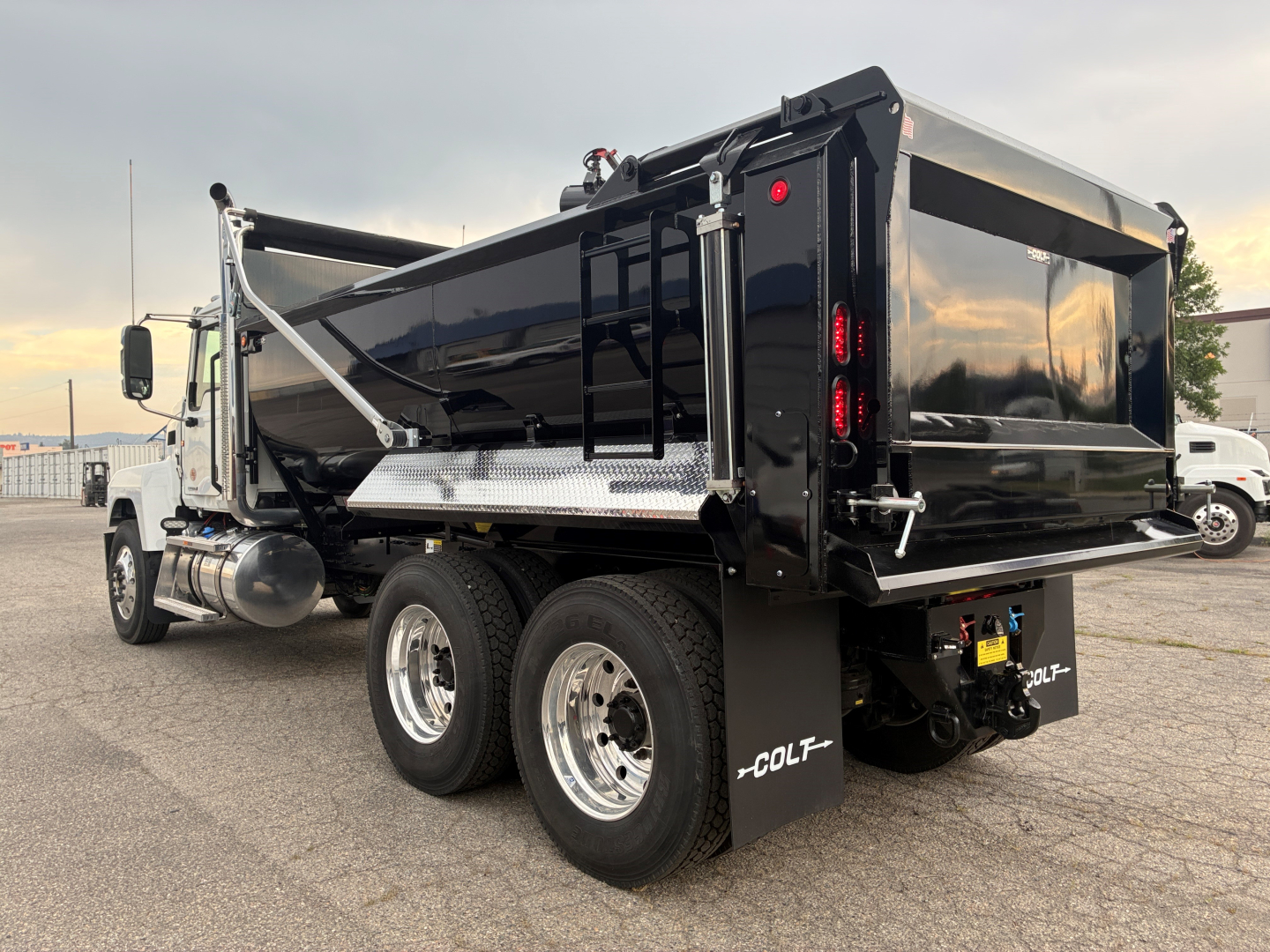 Truck rear/right side view showing a black dump body on a Mack PI64 chassis with dual rear axles, side step, and COLT branding on mud flaps; no visible text labels or numbers on body.