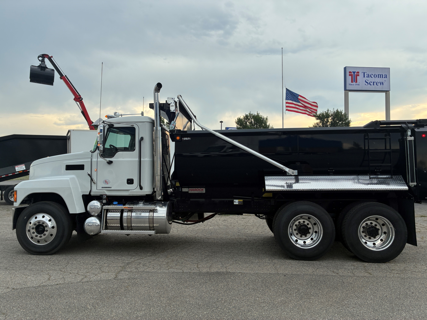 Mack PI64 dump truck side view with black dump body, white cab, chrome fuel tanks, and rear dual axle. visible text includes Mack emblem on grill and Tacoma Screw sign in background.