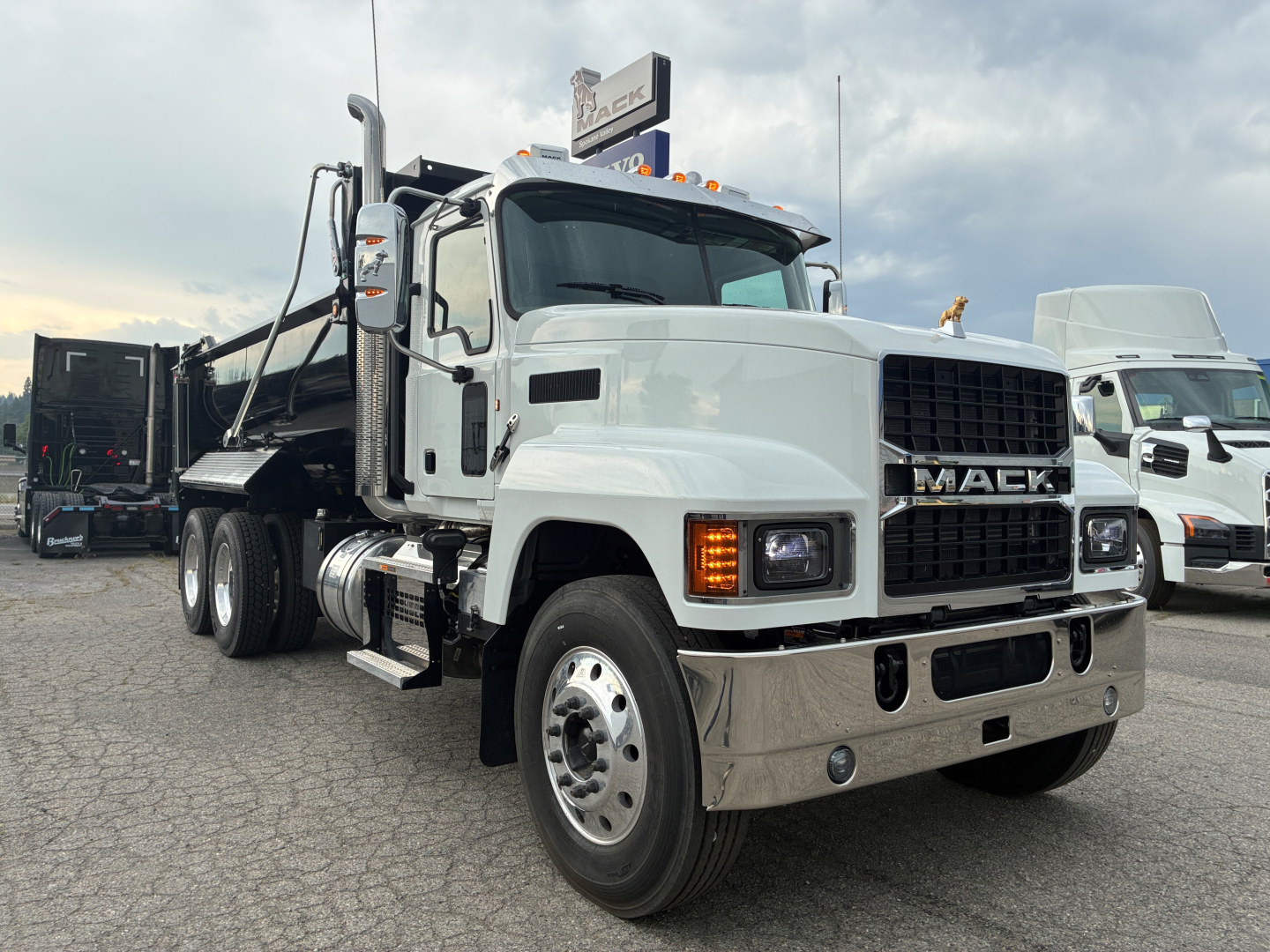 Dump truck front-left view showing white 2026 Mack PI64 with chrome bumper and grille, black dump bed, and dual rear axles; clear Mack logo on grille.