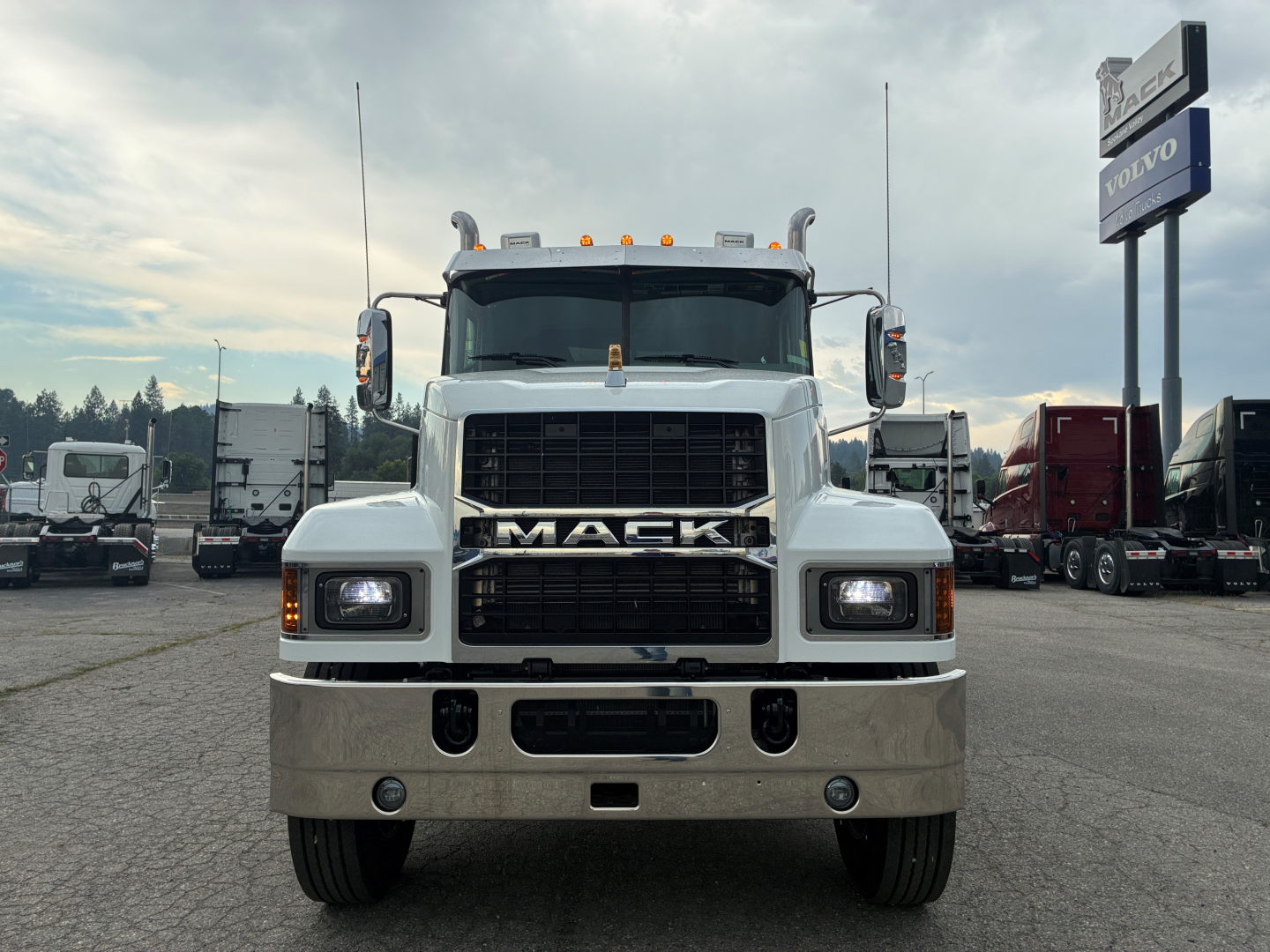 Truck front view showing Mack logo grille and large bumper with two round fog lights, plow-ready front, white cab, parking lot background.