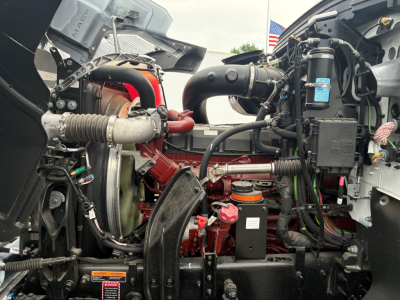 Engine bay of a 2026 Mack PI64 dump truck showing red engine, intercooler piping, hoses, wiring harness, and air intake components.