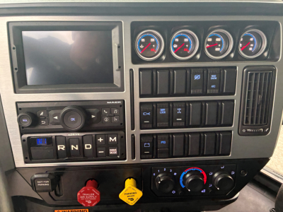 Dashboard view of a Mack PI64 cab interior showing gauges, infotainment screen, climate controls, and gear selector labeled R N D P M.