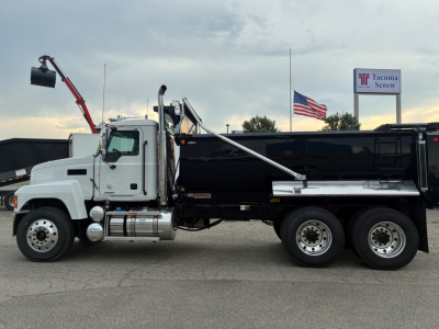 Mack PI64 dump truck side view with black dump body, white cab, chrome fuel tanks, and rear dual axle. visible text includes Mack emblem on grill and Tacoma Screw sign in background.