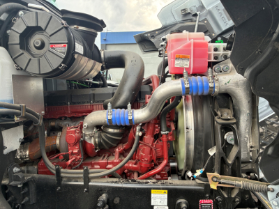 Mack PI64 engine bay with red diesel engine, intake piping, cooling fan, and pink coolant reservoir; visible hoses and components.