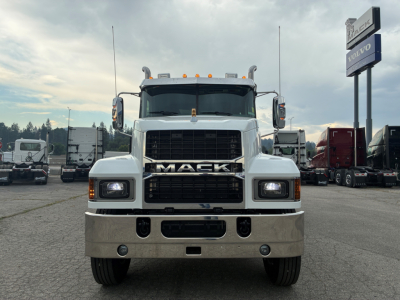 Truck front view showing Mack logo grille and large bumper with two round fog lights, plow-ready front, white cab, parking lot background.
