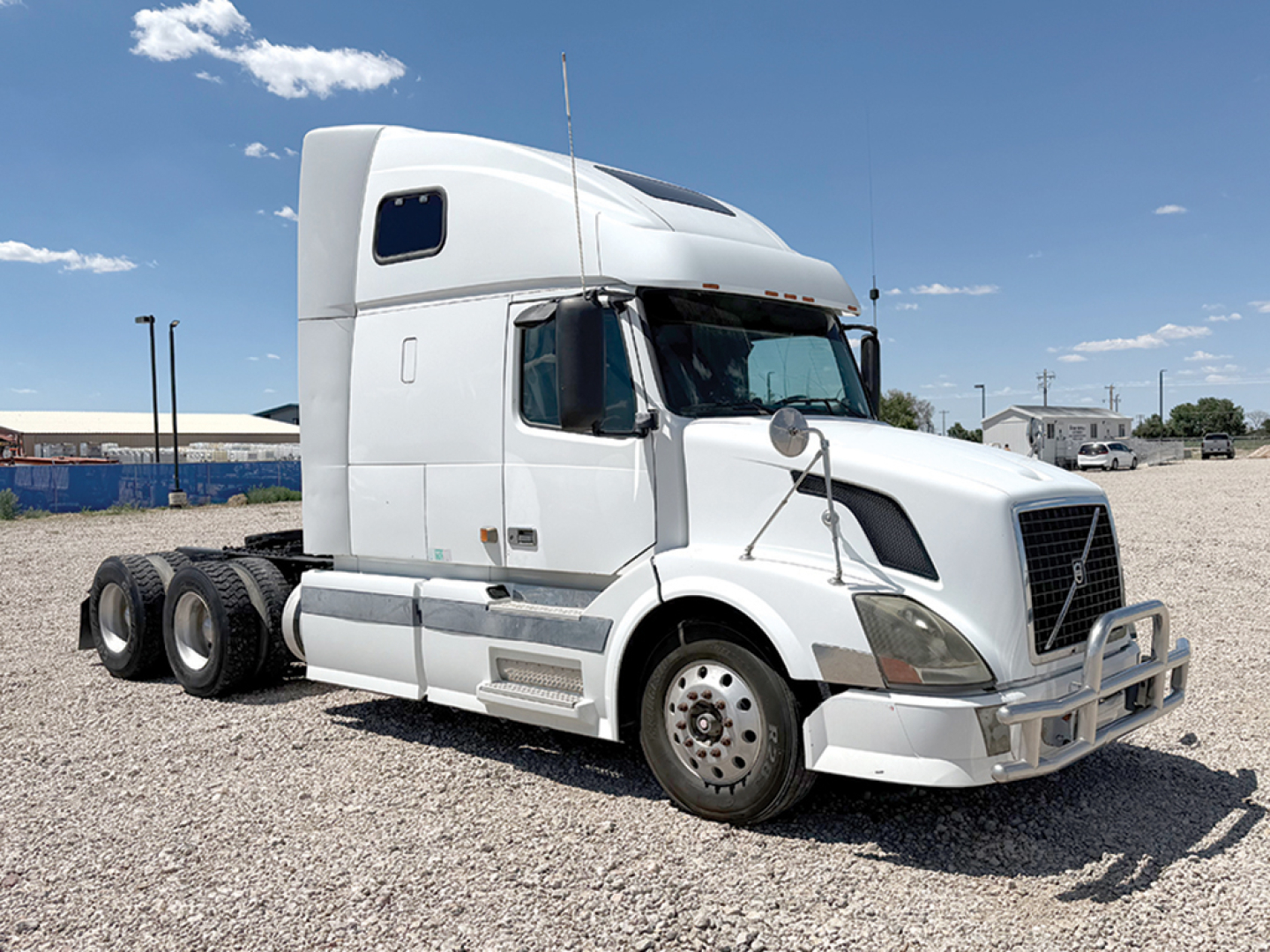 Truck cab showing Volvo VNL 670 sleeper with 6-wheeler rear axle configuration in gravel lot, white exterior, no visible branding beyond Volvo-inspired grille.