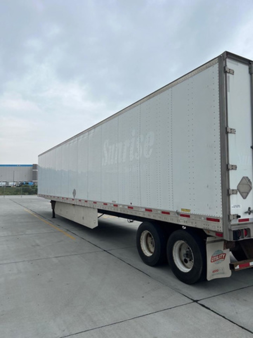 Trailer side view showing long box trailer with dual rear wheels and white exterior. Brand markings faint along side, rear doors visible.