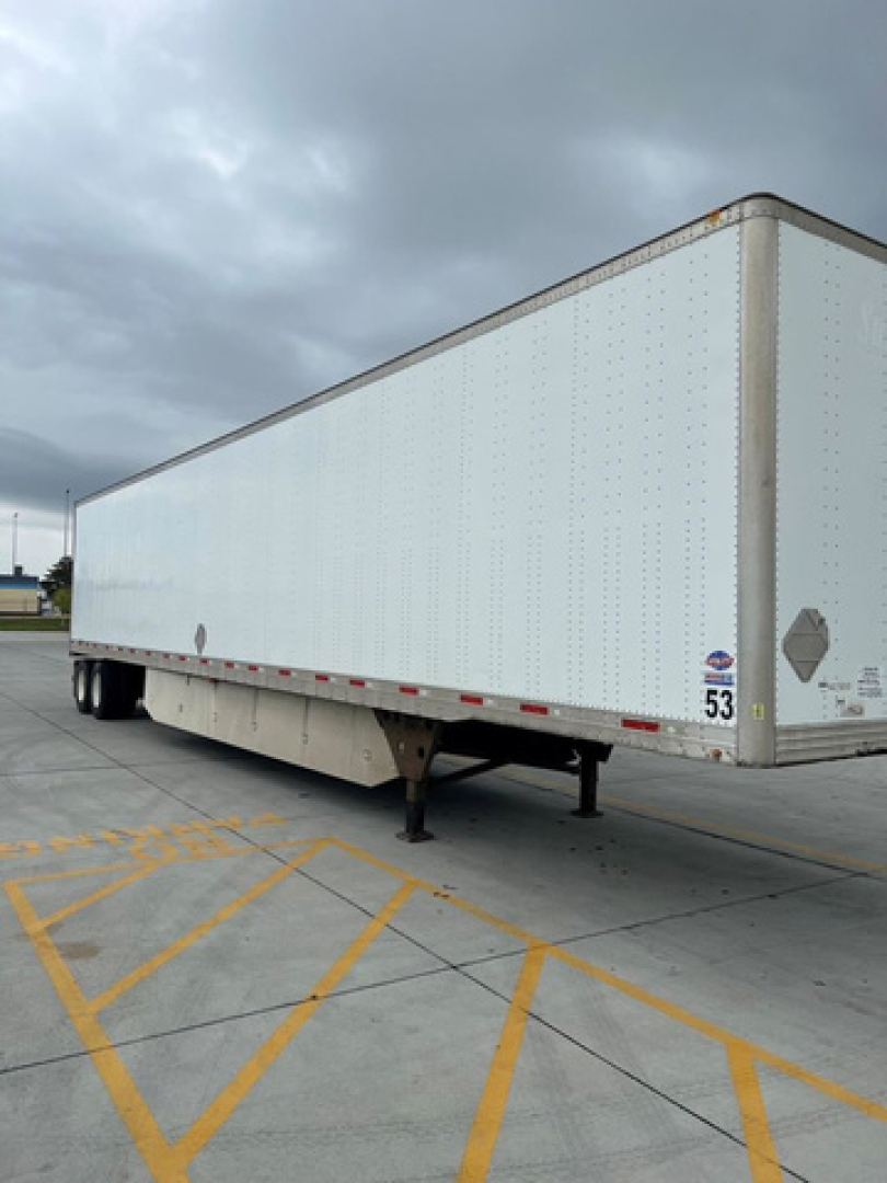 Utility van trailer showing long white box with wheels at the back and a support landing gear. DOT/brand stickers visible near the front corner.