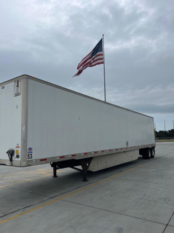Side view of a long white trailer with landing legs down, American flag on a flagpole, and a cloudy sky.