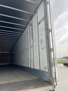 Trailer van interior showing cargo area with metal walls and open rear door; vertical corrugated walls and floor plate visible
