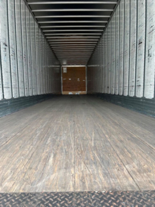 Trailer interior showing empty cargo space with wooden floor and metal sidewalls. End wall with rear doors visible.