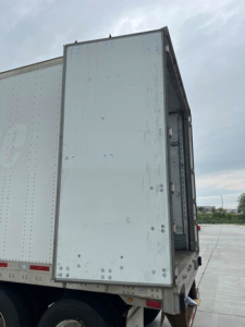 Rear open cargo trailer showing white metal doors and sidewall with rivets and hinges, utility van/trailer unit in background.