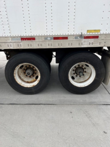 Trailer wheels and lower side of a utility van trailer showing dual rear axles with rims and tires, metal side panel, and reflective tape.