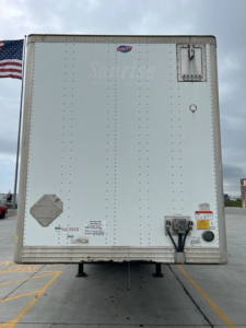 Rear view of a white utility van trailer with hitch and safety reflectors; some small labels near bottom left and yellow warning sticker; American flag in background