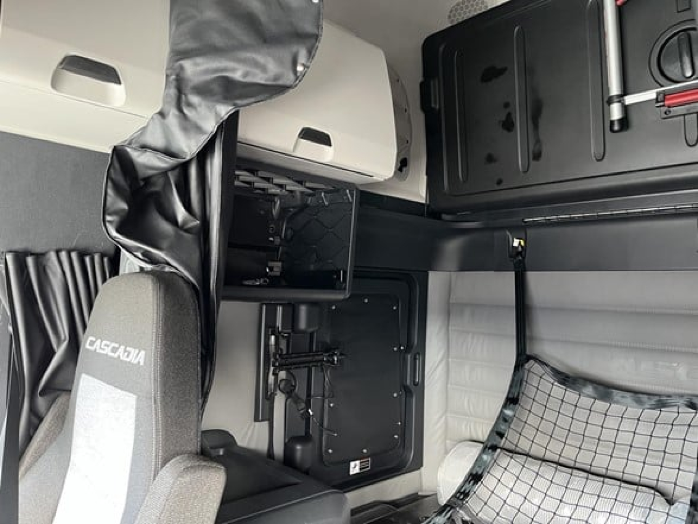 Truck interior of a 2026 Freightliner Cascadia 126, sleeper cab with driver seat, overhead storage, and dash; visible components include a storage cabinet, panel, and a chair labeled ‘CASCADIA’