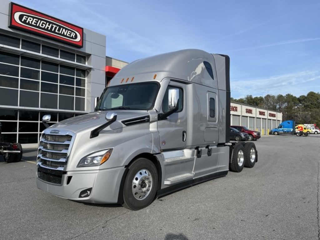 New 2026 Freightliner Cascadia 126 conventional sleeper truck in front of a dealership showing a chrome grille, aerodynamic body, and dual rear axles.