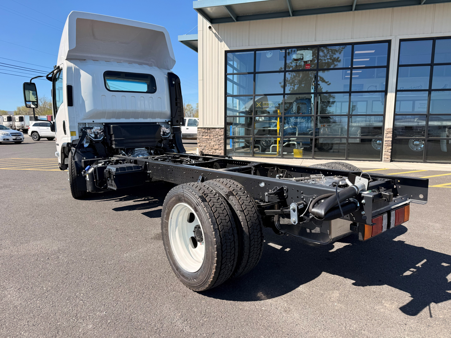 Isuzu NPRXD cab and chassis truck in showroom lot with open cab and chassis frame visible, front wheel, and dealership building in background.