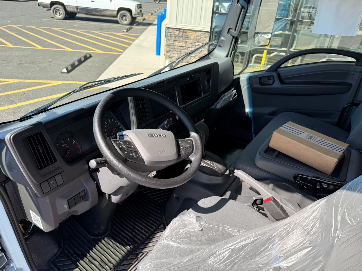 Isuzu NPRXD cab interior showing steering wheel with Isuzu logo, dashboard gauges, gear shifter, and a cardboard box on the passenger seat.