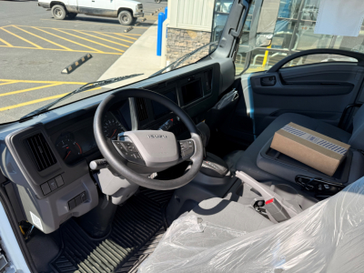 Isuzu NPRXD cab interior showing steering wheel with Isuzu logo, dashboard gauges, gear shifter, and a cardboard box on the passenger seat.