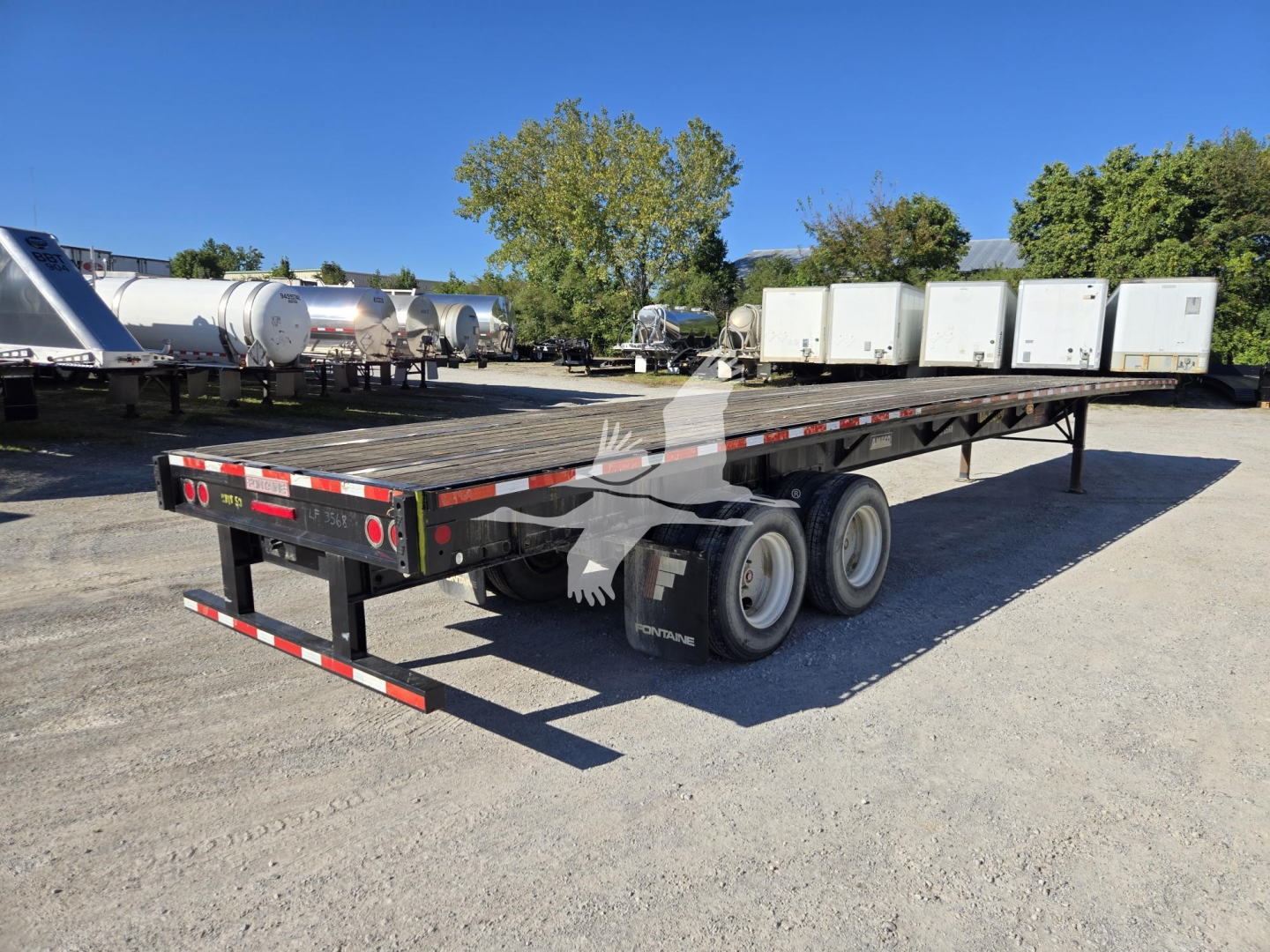 Flatbed trailer showing dual rear axles and long black deck with white and red reflective tape.