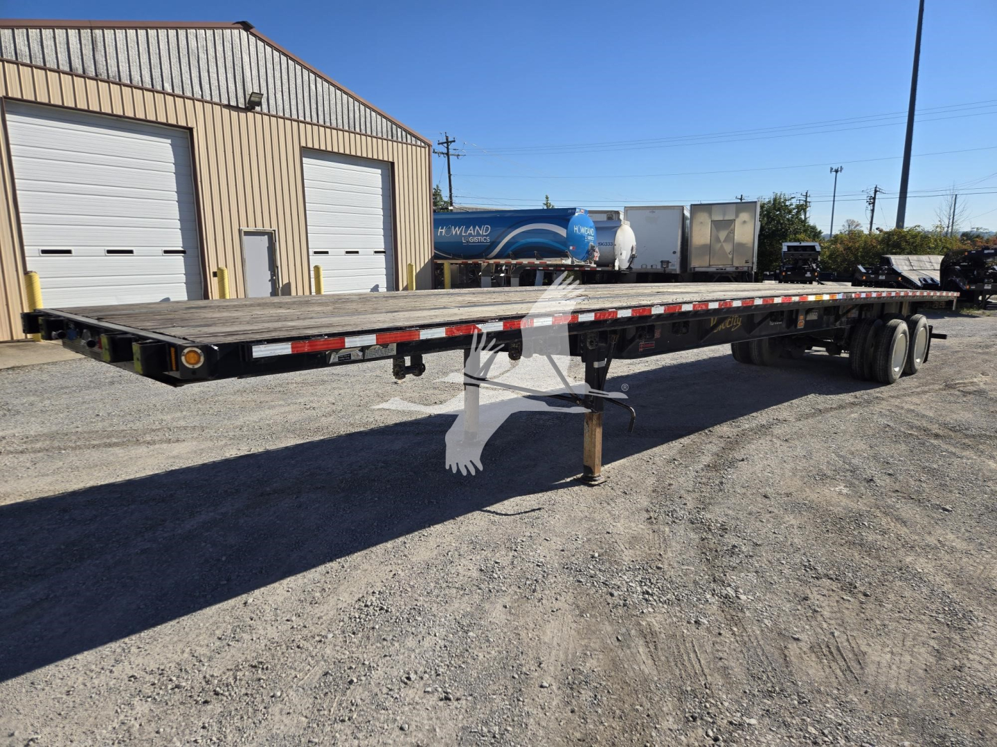 Flatbed trailer shown in side view with long deck and wheel axles at rear, support leg lowered, and industrial yard background.