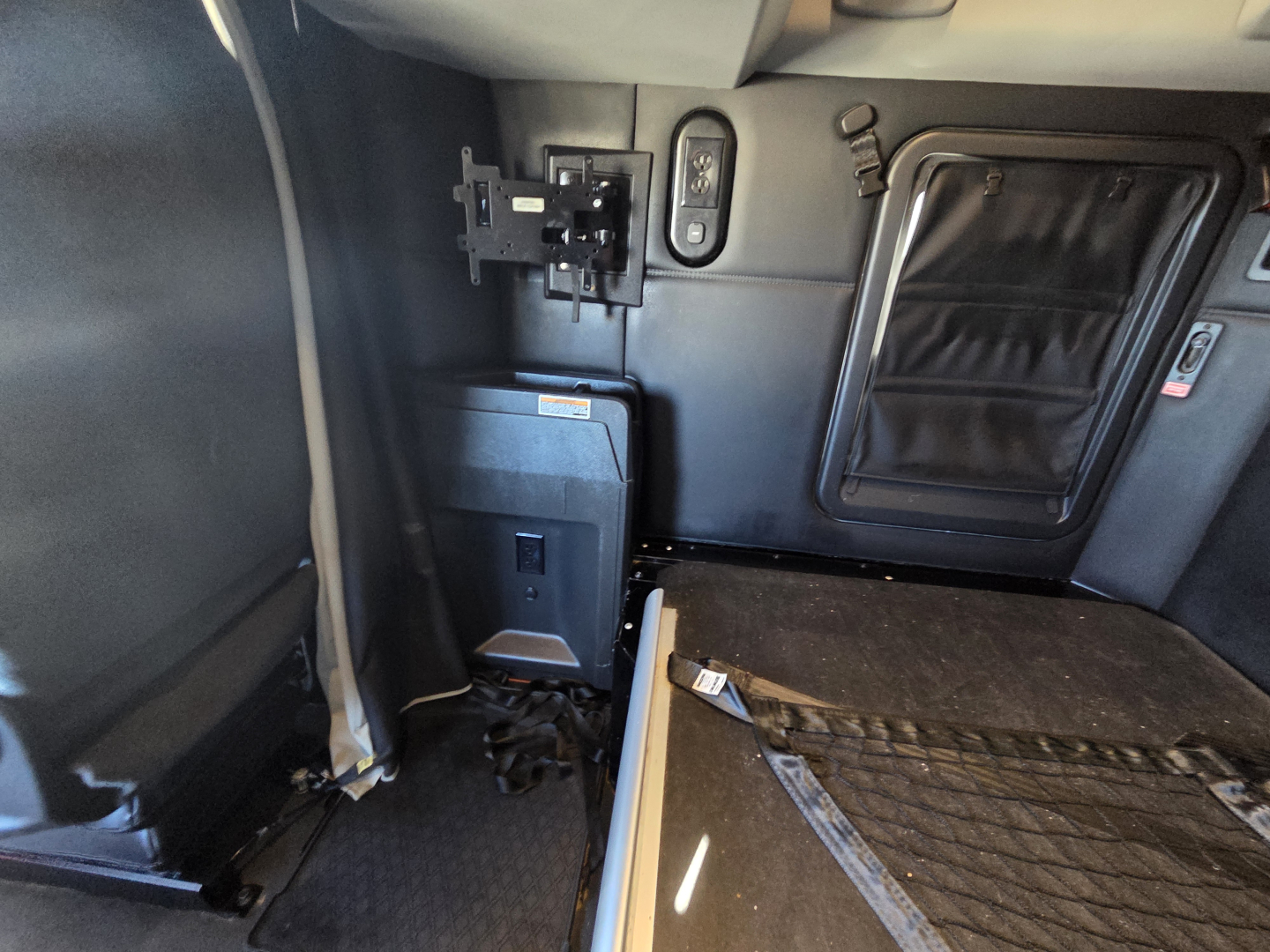 Truck cab interior showing control panel and mounting bracket with a small power box and a door, view from driver side with floor mat and bedding area.