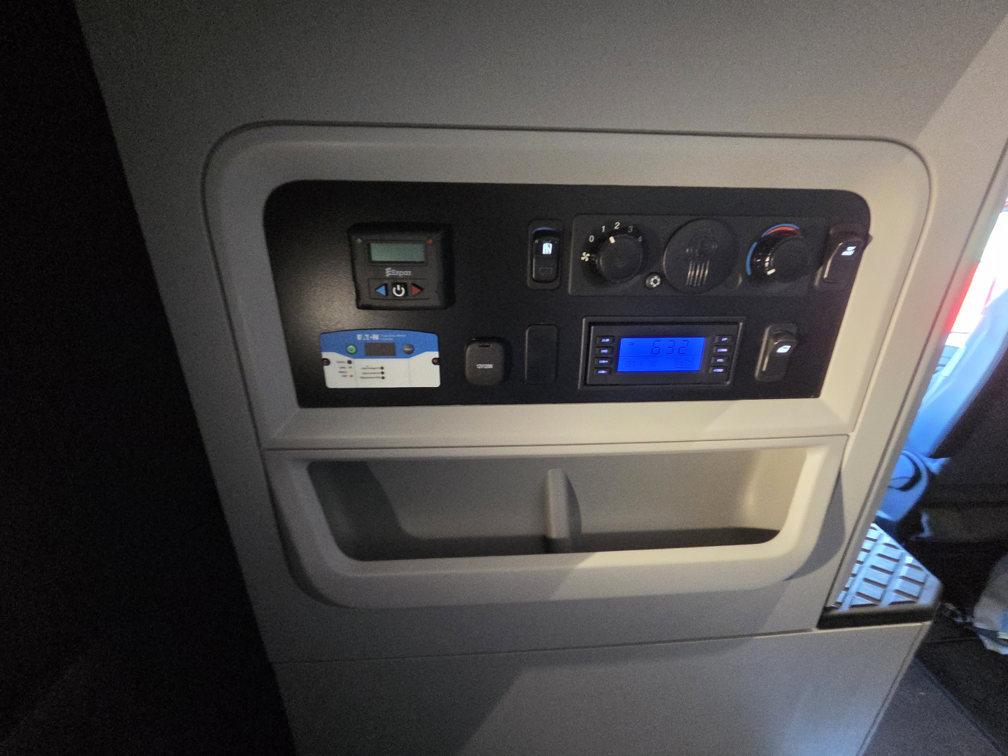 Kenworth engine or cab interior panel showing dashboard controls and a blue-lit display; includes knobs, digital readouts, and a small white card reader device.