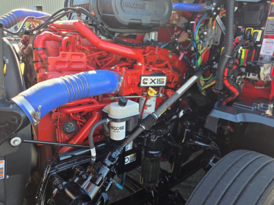 Kenworth T680 engine bay showing Cummins X15 engine with red cylinder head, blue turbo hose, ACCAR filter, and assorted wiring and fluid reservoirs.