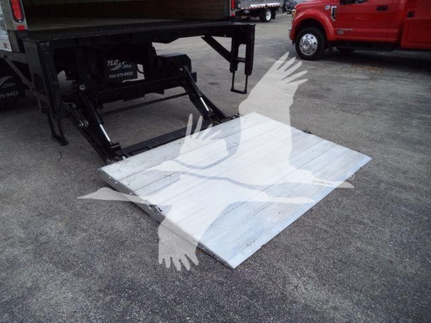Truck lift with a lowered aluminum loading ramp attached to a box truck tail; hydraulic legs and platform visible.