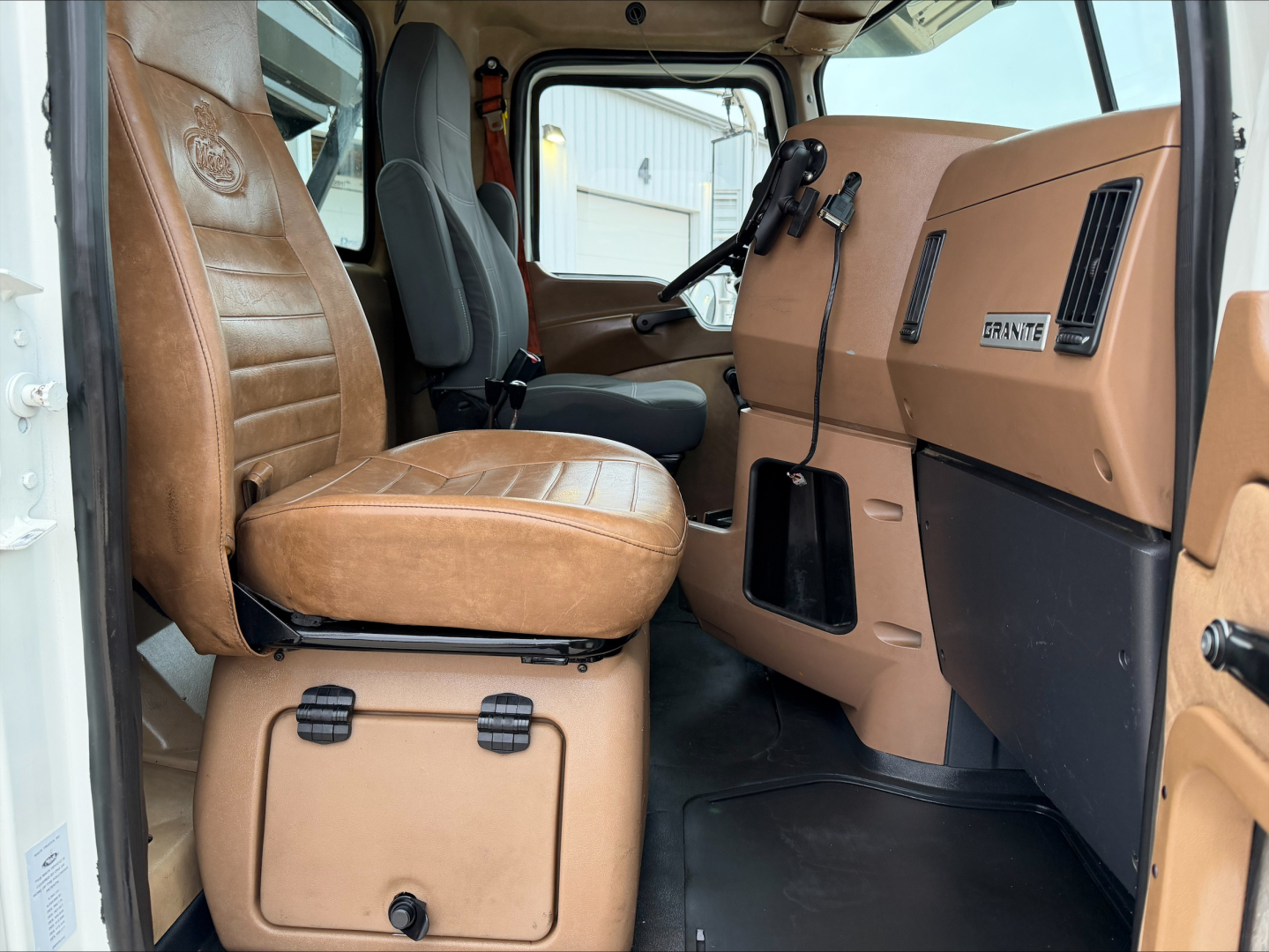 Cab interior of a Mack GU533 roll-off truck showing tan vinyl seats, dashboard with steering wheel, and door panels with basic controls.