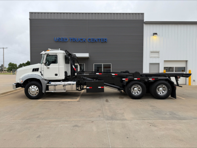 Side view of a Mack GU533 rolloff truck in front of a building labeled 'USED TRUCK CENTER' with a white cab and long black heavy-duty chassis.